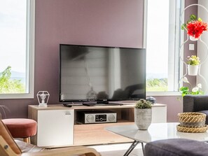 Ett modernt vardagsrum med en platt-TV, ett vitt skåp, ett litet bord med en växt och ett fönster med utsikt.