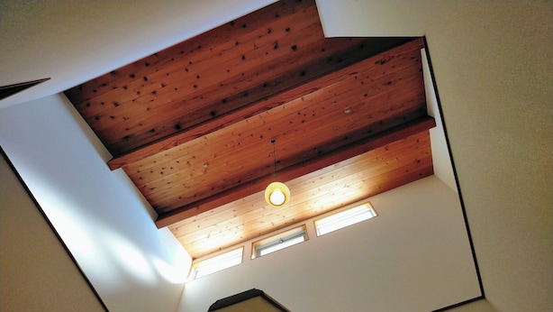 Techo inclinado con vigas de madera y una lámpara central. Dos pequeñas ventanas debajo de las vigas.