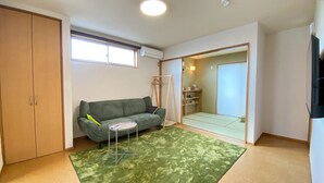 Una habitación con sofá verde, una mesita redonda pequeña, alfombra verde, armario de madera y una puerta corredera que da a otra habitación.