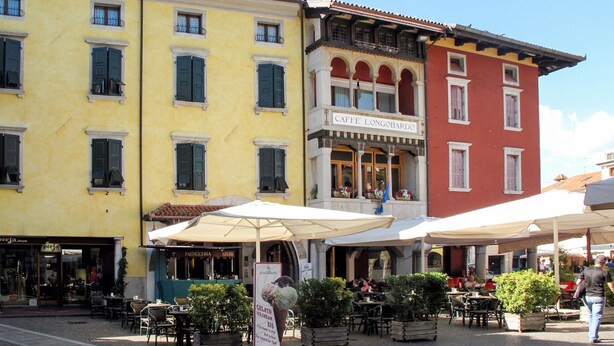 Une rue bordée de cafés avec des terrasses, un immeuble portant l’enseigne « CAFFE’ LONGOBARDO » et un magasin avec les enseignes « PASTICCERIA » et « GLAS ».