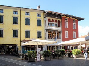 Una calle con mesas al aire libre en cafés, un edificio con el letrero 'CAFFE' LONGOBARDO, y una tienda con los rótulos 'PASTICCERIA' y 'GLAS'.