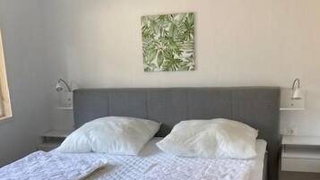 Un dormitorio con una cama gris, ropa de cama blanca, una mesita de noche pequeña con una lámpara y una planta en la pared.