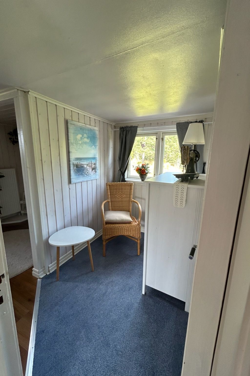 Een kamer met een rieten stoel, een tafeltje, een schilderij, een lamp en een raam met gordijnen.