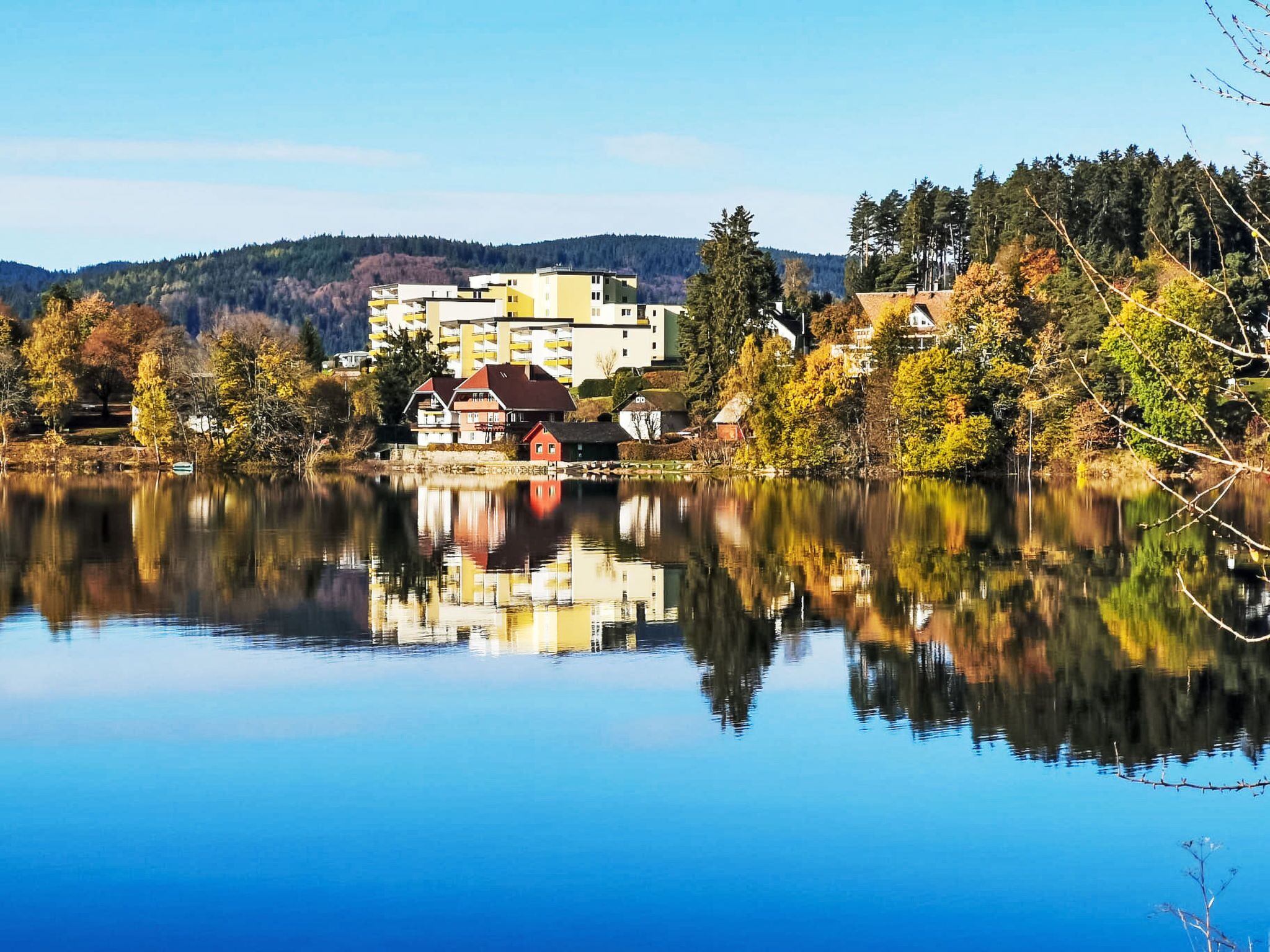 Ein ruhiger See, auf dessen Oberfläche Gebäude sich spiegeln, umgeben von Bäumen und Hügeln.