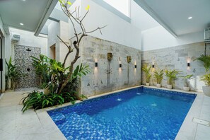 An indoor swimming pool with a tiled floor, surrounded by potted plants and a stone wall with decorative elements.