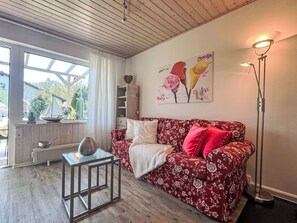 Een woonkamer met een houten plafond, een rode bloemenbank, een glazen salontafel, een vloerlamp en een bloemschilderij.
