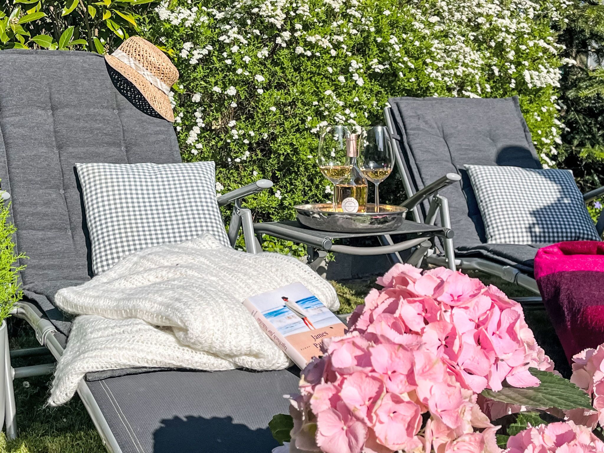 Twee lounge stoelen met geruit kussens, een geweven hoed, een dienblad met glazen en een fles, en roze bloemen.