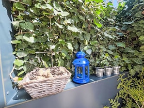 Ein blauer Laternen, ein Korbwannen und Metallbehälter auf einem Balkon mit grünen Blättern im Hintergrund.