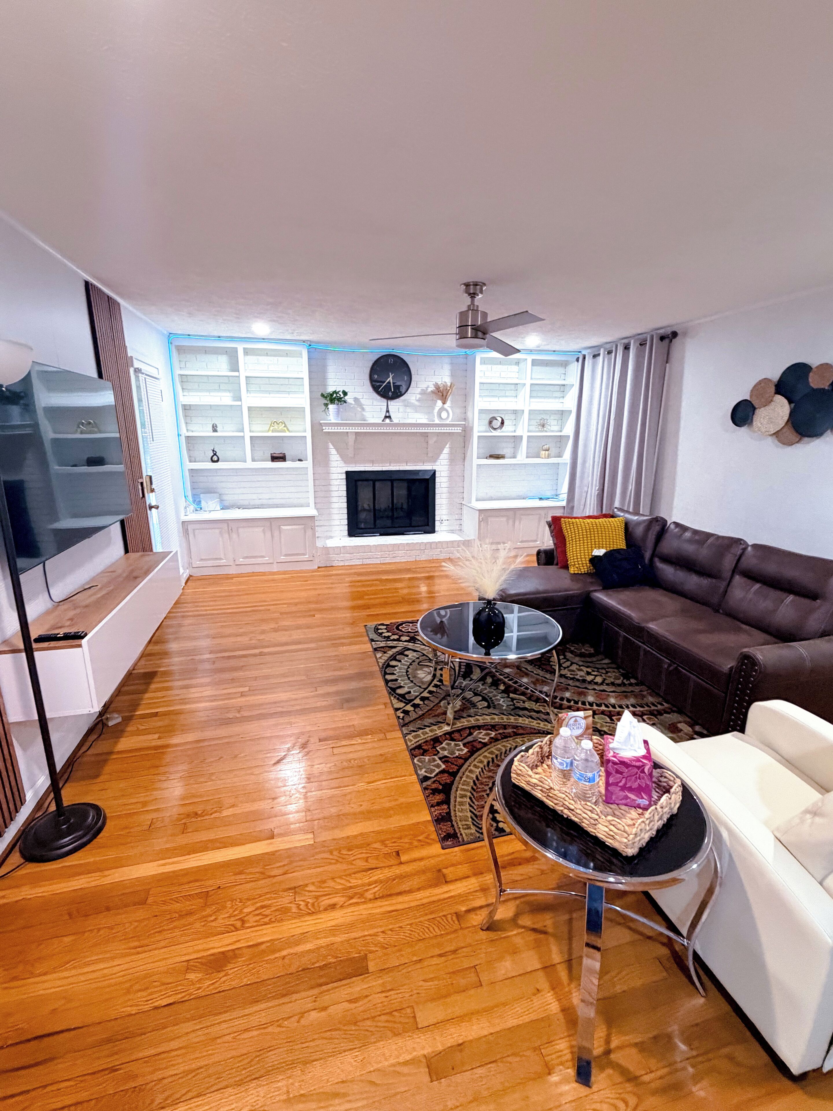 A living room with a fireplace, a brown leather sofa, a white armchair, a coffee table, and a TV on a stand.
