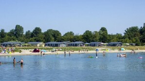 En semesteranläggning vid en sjö med flera stugor, en sandstrand och människor som ägnar sig åt olika vattenaktiviteter.