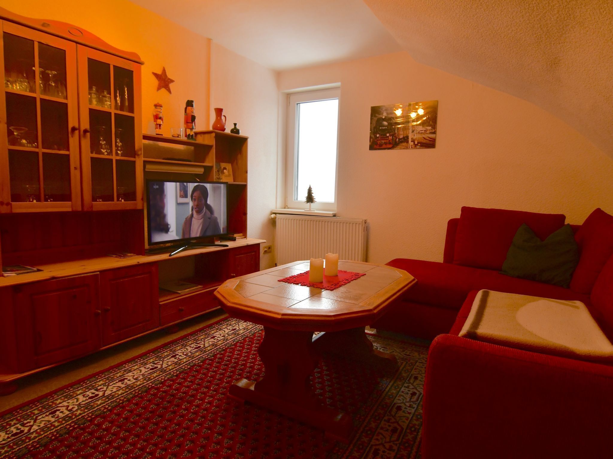 A cozy living room with a television, red furniture, a patterned rug, and a window with a view.