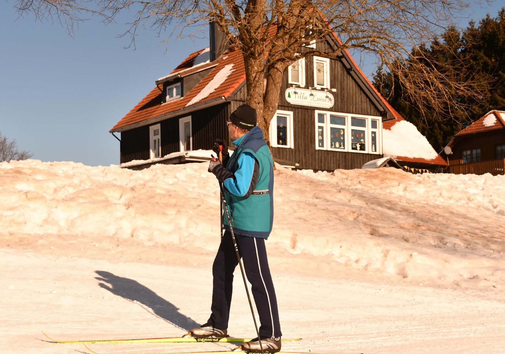 Une personne fait du ski sur une pente enneigée, avec une maison en bois en arrière-plan.