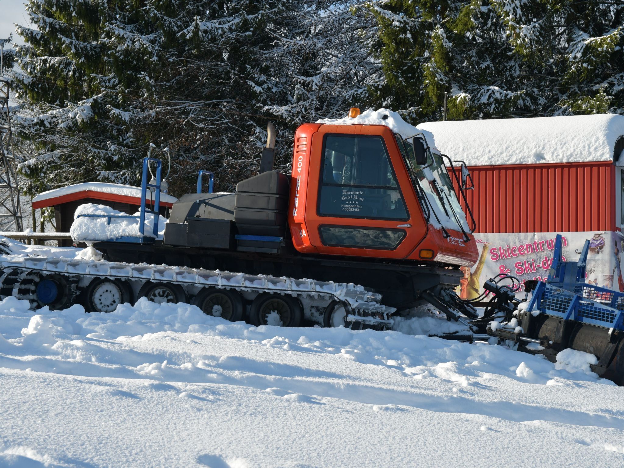 A snowplow truck parked in a snowy area with a red building in the background.