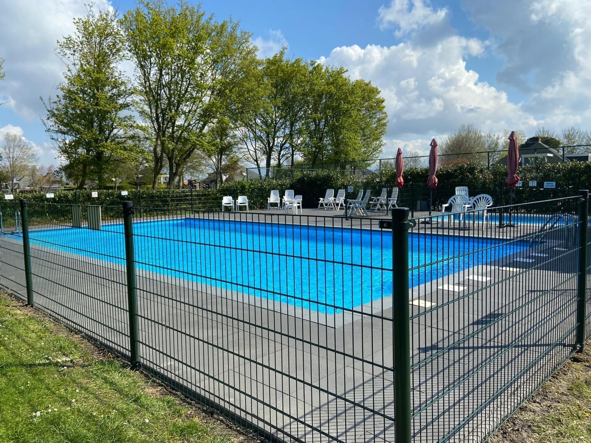 Ein rechteckiger Pool, umgeben von einem schwarzen Metallzaun, mit weißen Liegestühlen und roten Sonnenschirmen am Rand.