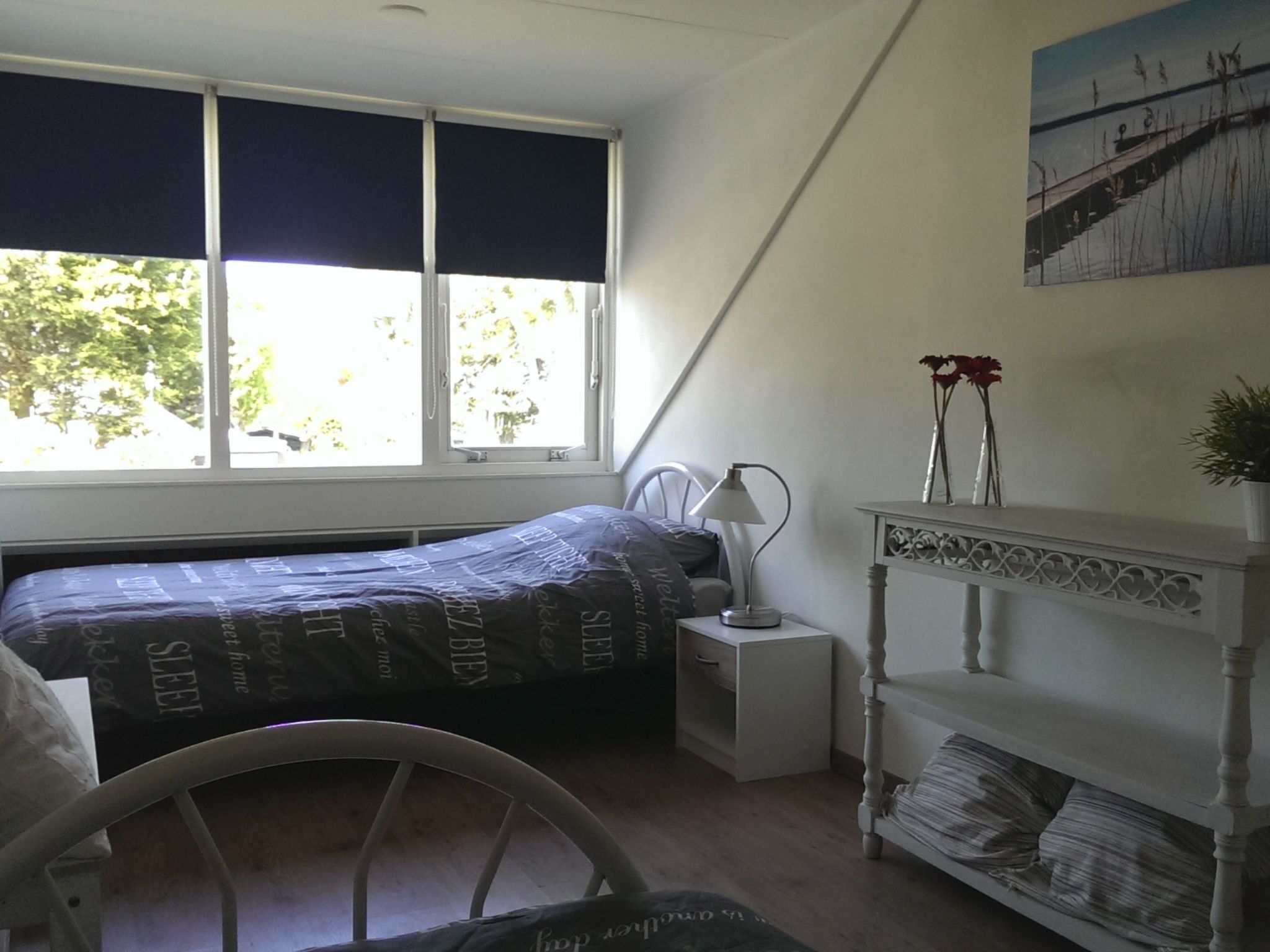 Een slaapkamer met een bed, nachtkastje, raam met jaloezieën en een schilderij aan de muur.