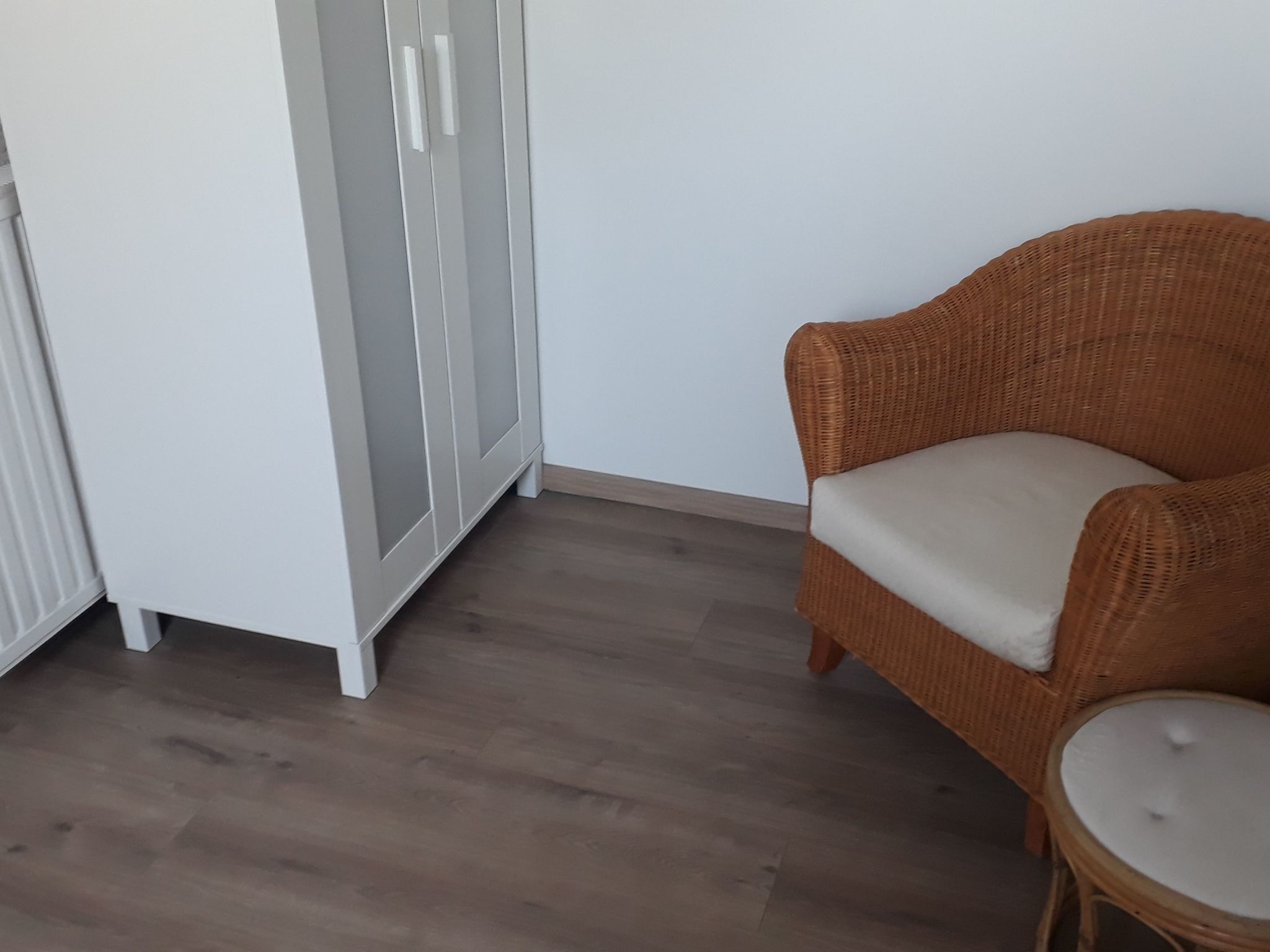 A room with a wicker chair and a small round table, a white wardrobe, and a wooden floor.