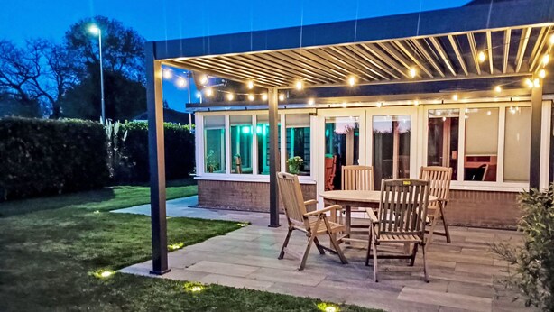 Eine Terrasss mit Tisch und Stühlen, ein überdachter Sitzbereich mit Lichterketten und ein Hecken im Hintergrund.