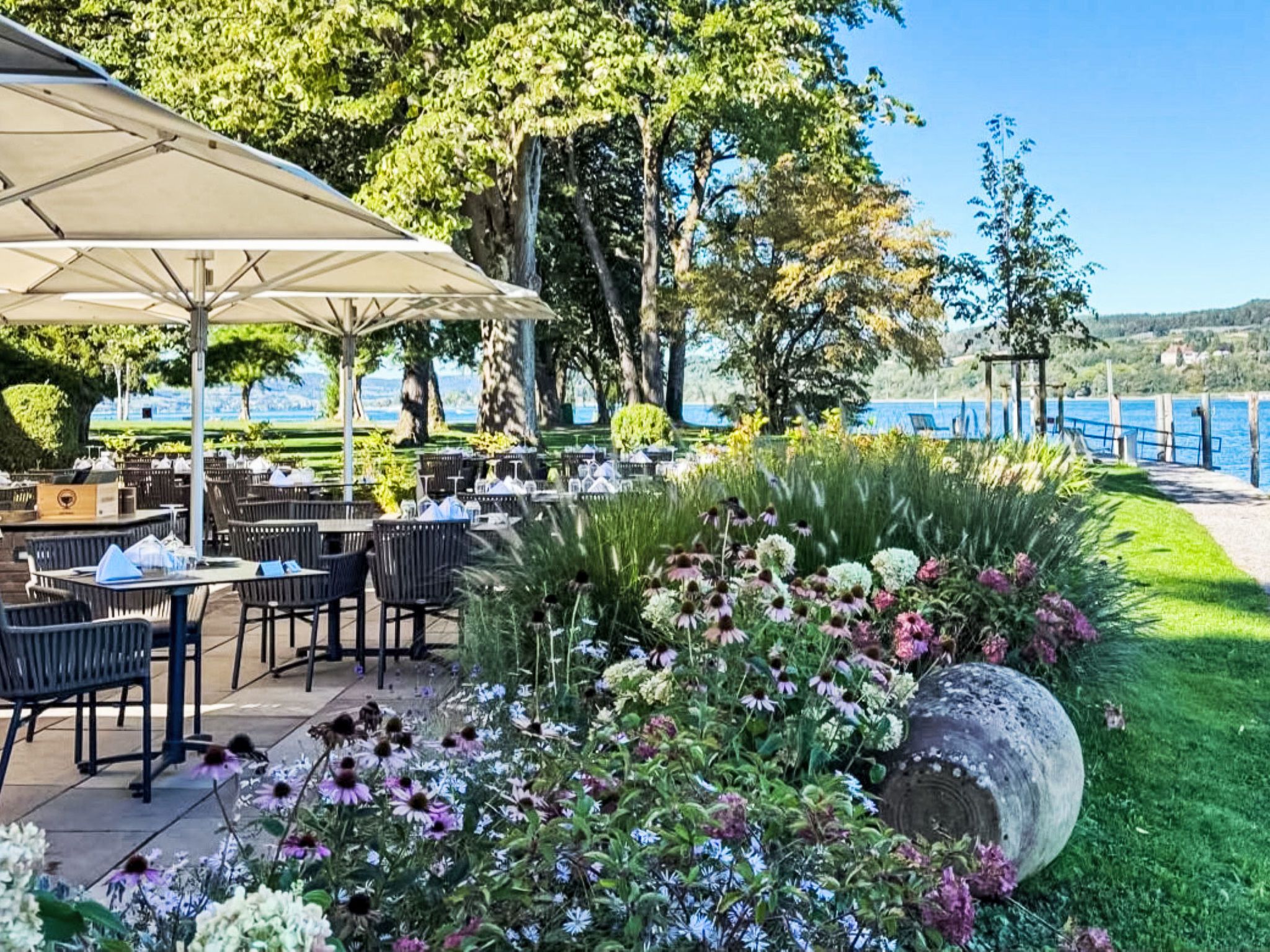 Ein Essbereich im Freien mit Tischen und Stühlen, umgeben von einem Garten mit verschiedenen Blumen und Blick auf einen See.