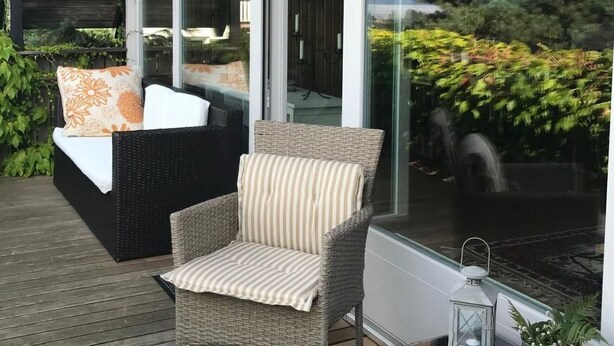 Een terras buiten met rieten meubilair, een glazen deur naar binnen en uitzicht op groen.