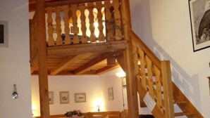 A wooden staircase leading to a lofted area with a balcony, a dining area with a table and chairs, and a floral-patterned ottoman.