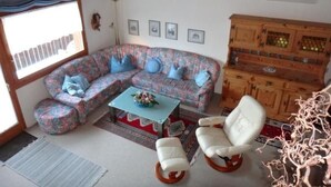 A cozy living room with a floral sofa, a small coffee table, and a white armchair. There is a wooden cabinet with glass doors and a window with blinds.