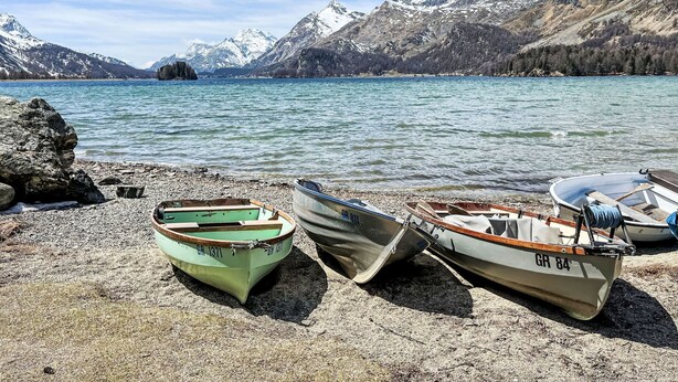 Three boats on a rocky shore by a lake with mountains in the background.