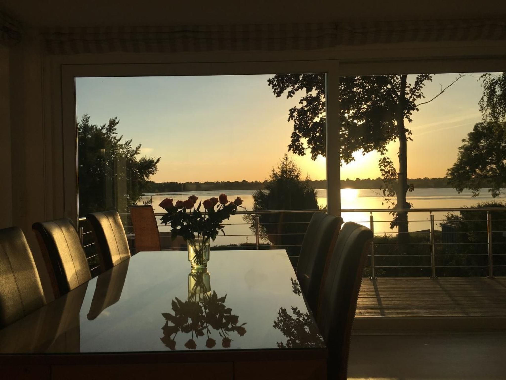 A dining table with chairs, a vase with flowers, and a view of a lake at sunset.