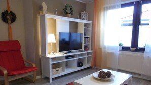 Ein Wohnzimmer mit Fernseher, einem roten Sessel, einem weißen Couchtisch und einem fenster mit Vorhängen.