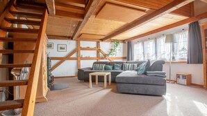 Ein gemütlicher Loftraum mit freiliegenden Holzbalken, einem grauen Ecksofa, einem kleinen Holztisch und einer Treppe, die zu einer erhöhten Ebene führt.