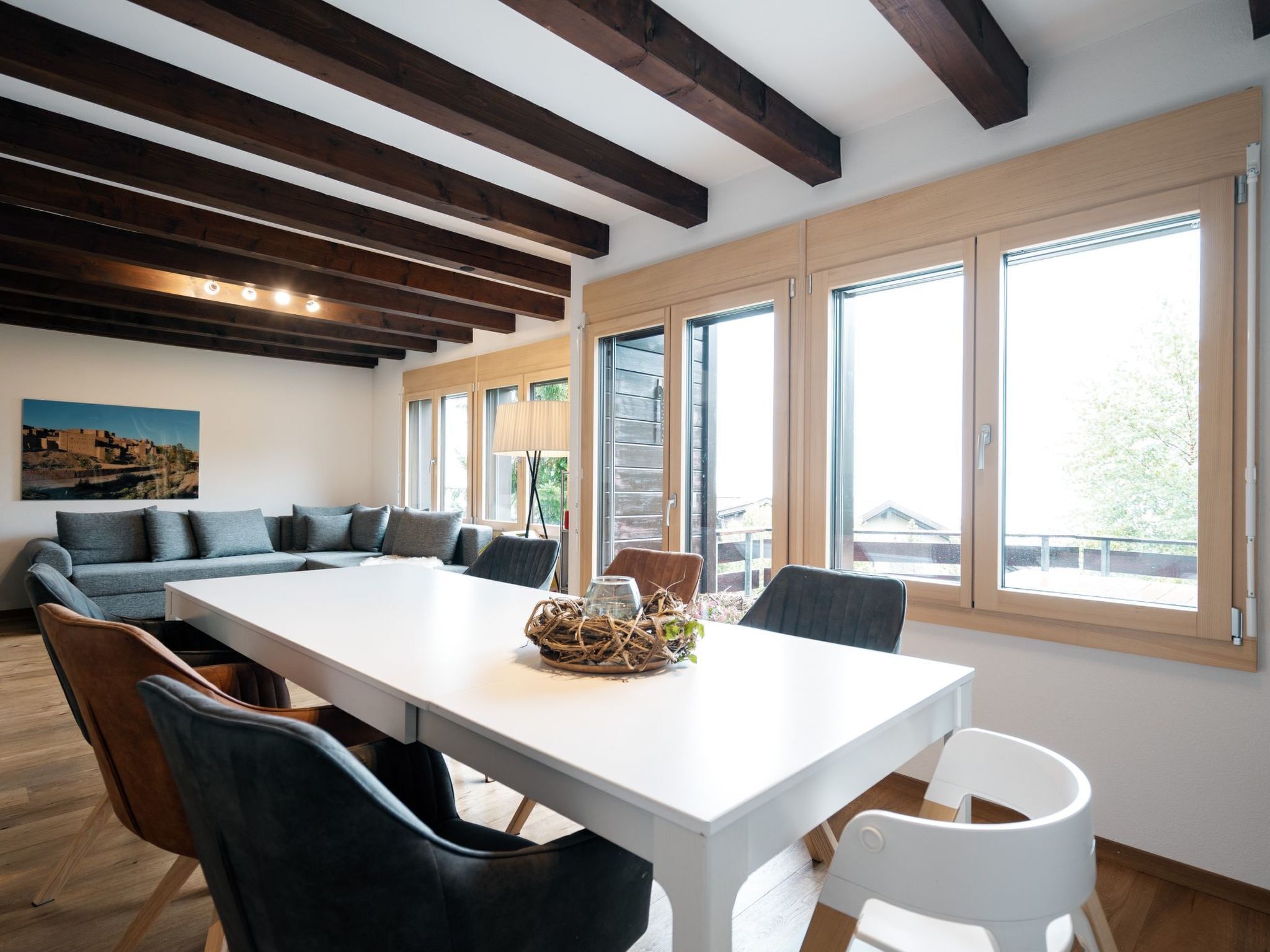 Ein modernes Wohnzimmer mit einem großen weißen Tisch, dunklen Sofas und Blick ins Freie durch große Fenster.