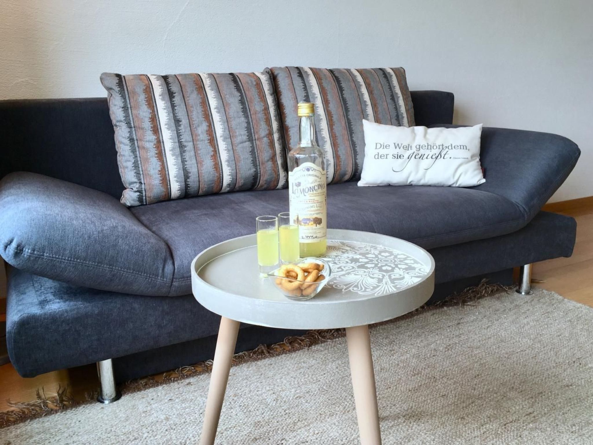 A modern living room with a grey sofa, striped pillows, a round coffee table with a bottle of liquor, glasses, and snacks.