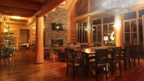 A cozy log cabin interior with a fireplace, wooden dining table, and large windows.