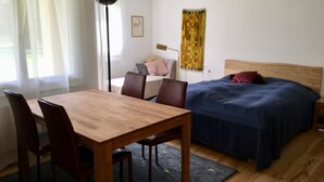 Ein Zimmer mit Holztisch und Stühlen, einem Bett mit blauer Bettdecke, einer Stehlampe und einem Fenster mit durchsichtigen Vorhängen.