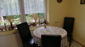 A room with a round table covered with a white lace tablecloth, two black chairs, a radiator, and a window with lace curtains. - Landblick by Interhome (Friederikensiel)