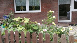 A brick house with two windows, a wooden fence, and a garden with various plants and flowers. - Landblick by Interhome (Friederikensiel)