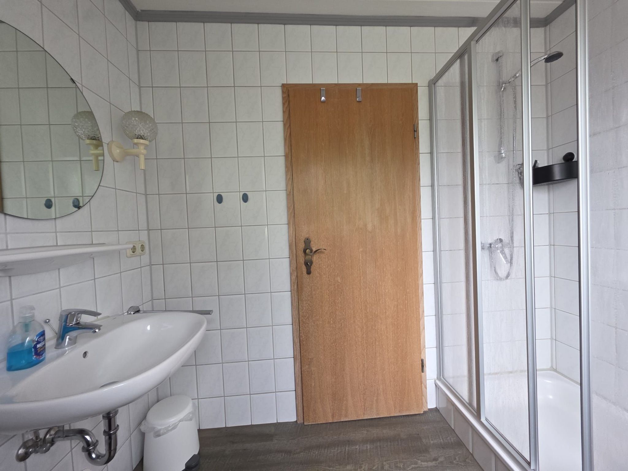 A bathroom with white tiled walls, a wooden door, a white sink with a mirror above it, and a shower area.