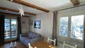Um quarto acolhedor com teto de madeira, um sofá cinza, uma mesa de jantar de madeira com cadeiras e janelas amplas com vista para árvores cobertas de neve.