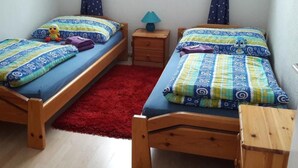 Un dormitorio pequeño con dos camas de madera, cada una con ropa de cama estampada y un peluche. Una alfombra roja separa las camas, y hay una mesita de noche con una lámpara entre ellas.