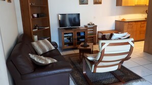 Una sala de estar con un sofá de cuero marrón, un mueble bajo de televisión de madera, una mesita pequeña de madera y una zona de cocina con armarios de madera.