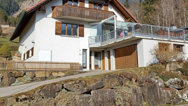 Un chalet de dos pisos con un balcón, un porche cubierto y un muro de piedra de contención al frente.