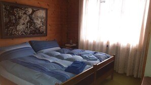 Puinen huone, jossa on sänky, yöpöytä, seinällä taulu ja ikkunassa verhot.