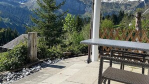 Eine Terrasse mit Steintisch und Korbstühlen, mit Blick auf eine gebirgige Landschaft mit schneebedeckten Gipfeln.