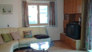 En stue med en beige sofa, et glasbord, et fjernsyn på et træskab og et vindue med gardiner.