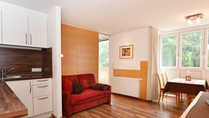 Ett kompakt bostadsutrymme med ett litet kök, en röd soffa, ett matbord med stolar och ett stort fönster med utsikt över grönområden.