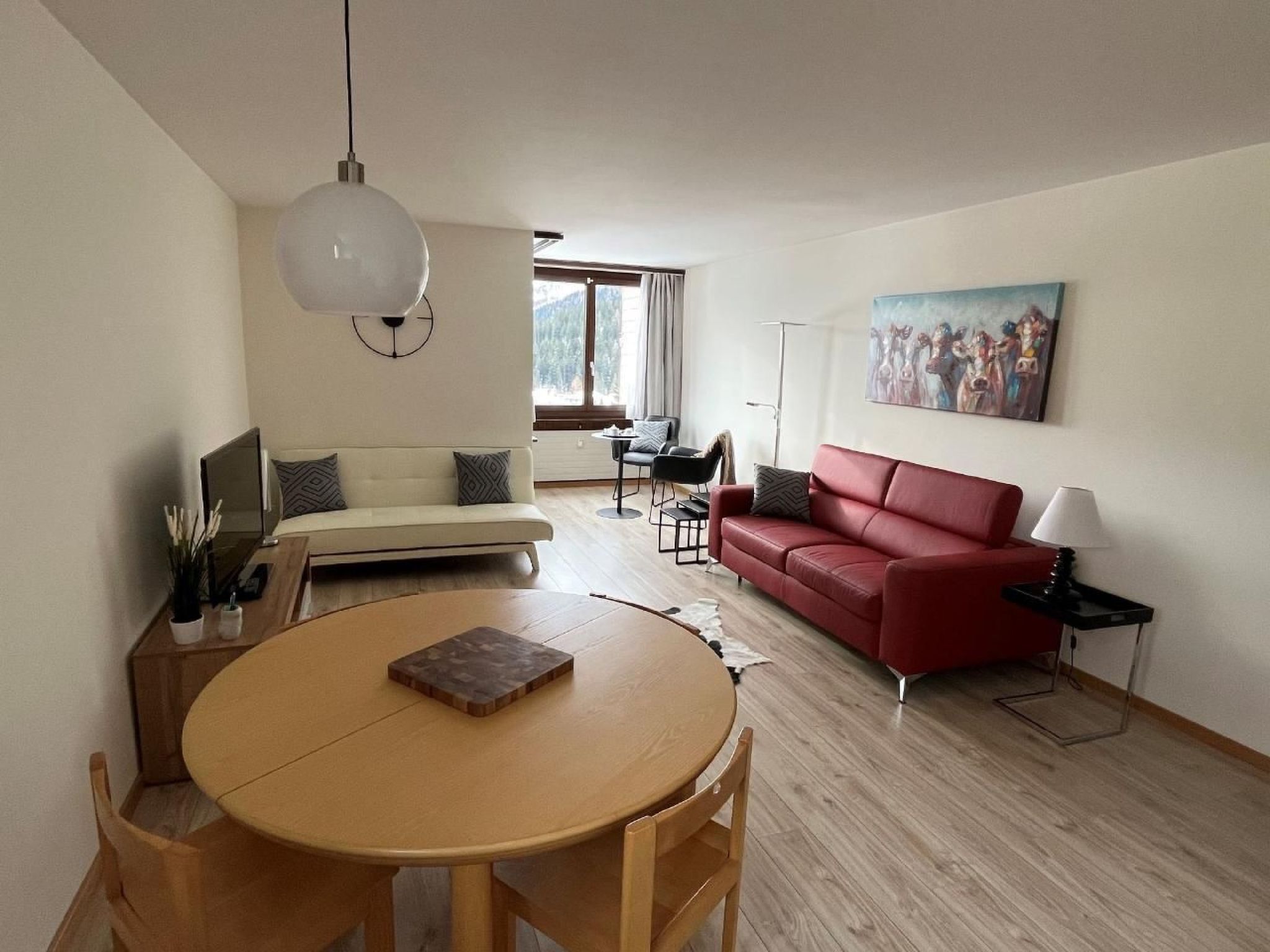 Een moderne woonkamer met een ronde houten tafel, een rode bank, een witte bank en een schilderij aan de muur.