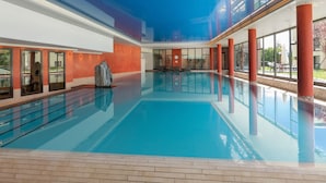 En stor indendørs swimmingpool med krystalklart blåt vand, omgivet af glasvægge og røde søjler.