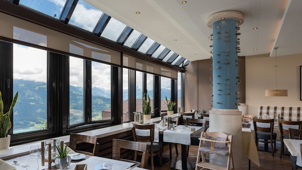 En restaurant med store vinduer der giver udsigt til bjergene, træborde og stole, og en blå søjle i midten.