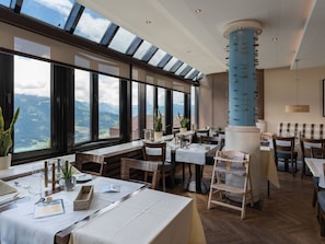 Ein Restaurant mit großen Fenstern und Blick auf die Berge, Holztische und -stühle sowie eine blaue Säule in der Mitte.