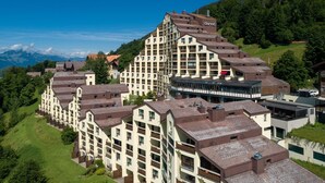 Et moderne hotelkompleks bestående af flere bygninger, herunder et markant tårn med balkoner, beliggende i et bjergrigt område med frodig vegetation og blå himmel.