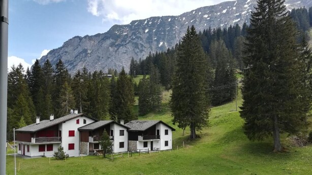 Une rangée de maisons modernes, aux façades blanches rehaussées d’éléments rouges, se dressant sur un paysage montagneux et bordée d’une forêt dense.